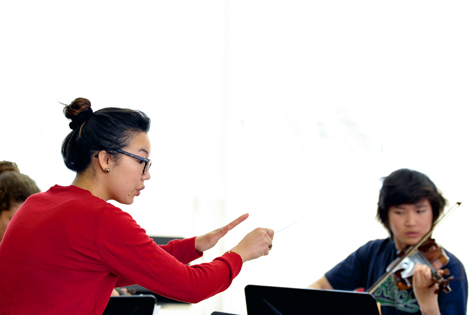 An Asian female conductor, teaching a male student, who is playing the violin.